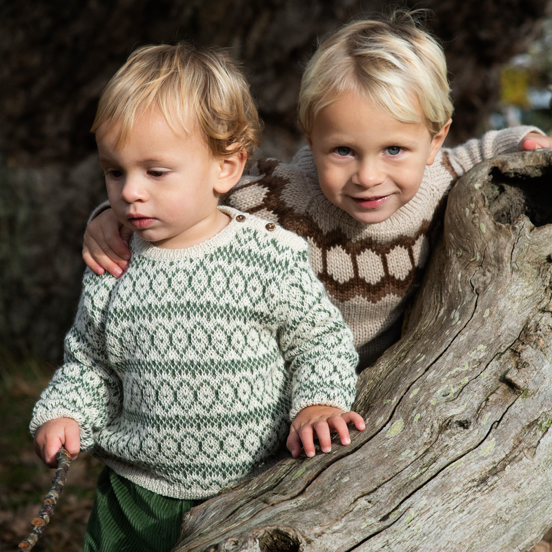 Two young children wearing knit Fair Isle sweaters posed outdoors next to a large weathered log. The child in front wears an off-white sweater with green geometric patterns, and the child behind wears a beige sweater with brown geometric patterns.
