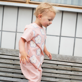 Child wearing a pink floral outfit standing on a wooden bench outdoors.