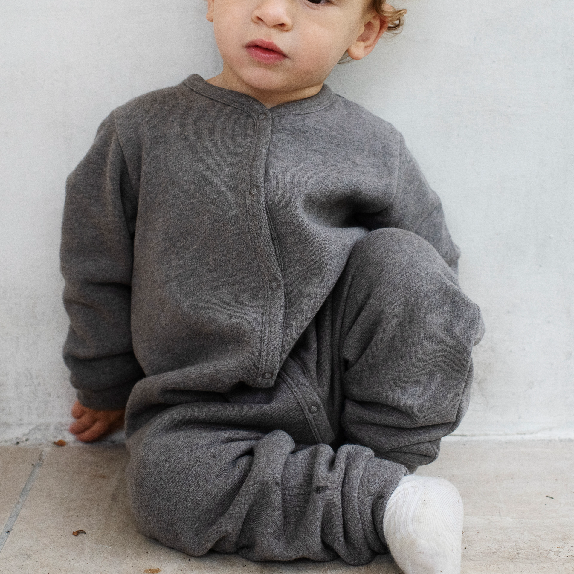 A baby wearing a dark gray brown heathered fleece sleeper/jumpsuit with a snap-button front, sitting against a light gray concrete wall.