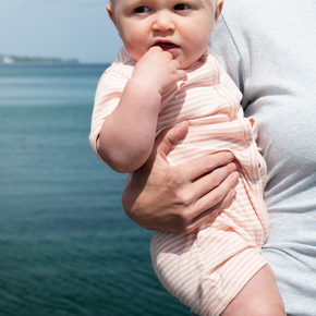 Baby in a pink outfit being held by an adult with a body of water in the background