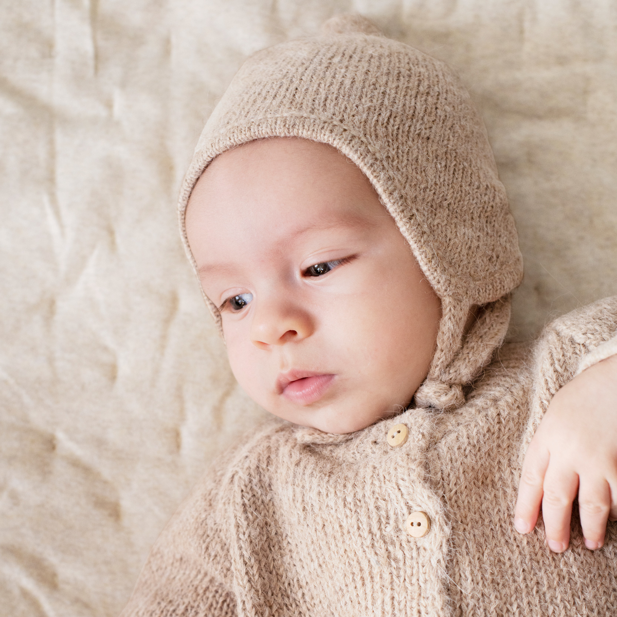 Baby wearing a beige knit textured cardigan and matching bonnet on a neutral background.