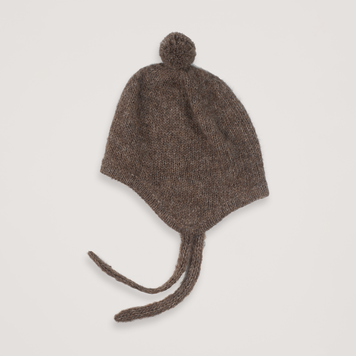 Brown knit baby bonnet with a pompom and ties on a neutral background.