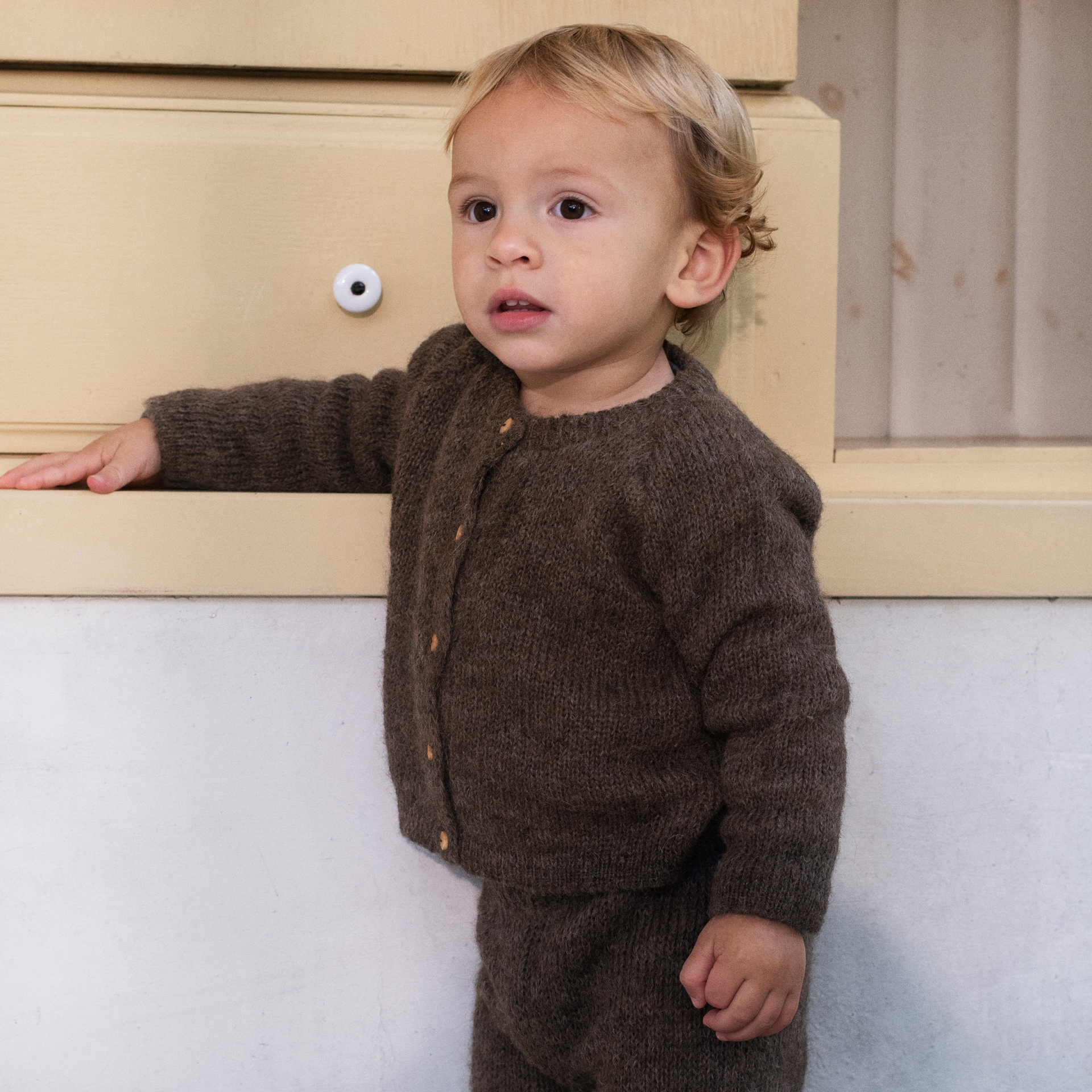 A toddler standing outdoors against a light wall, wearing a brown knit cardigan and matching pants.