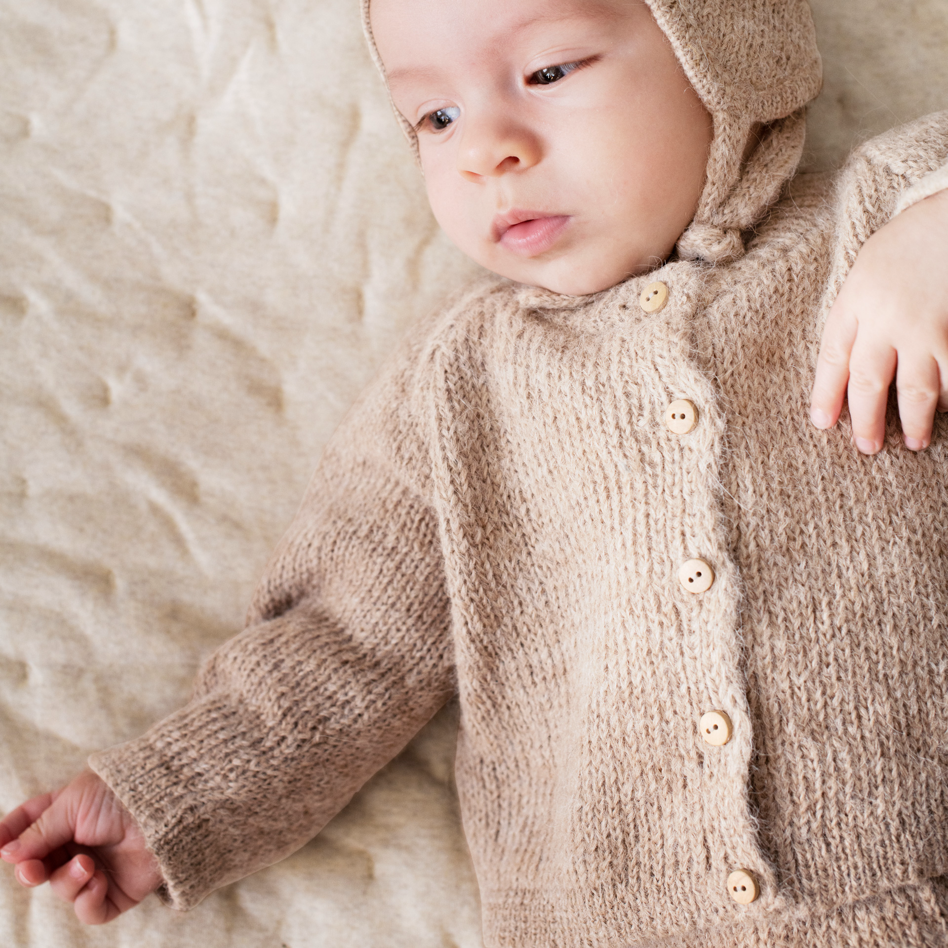 A close-up of a baby wearing a beige knit cardigan and matching bonnet, lying on a textured beige blanket.