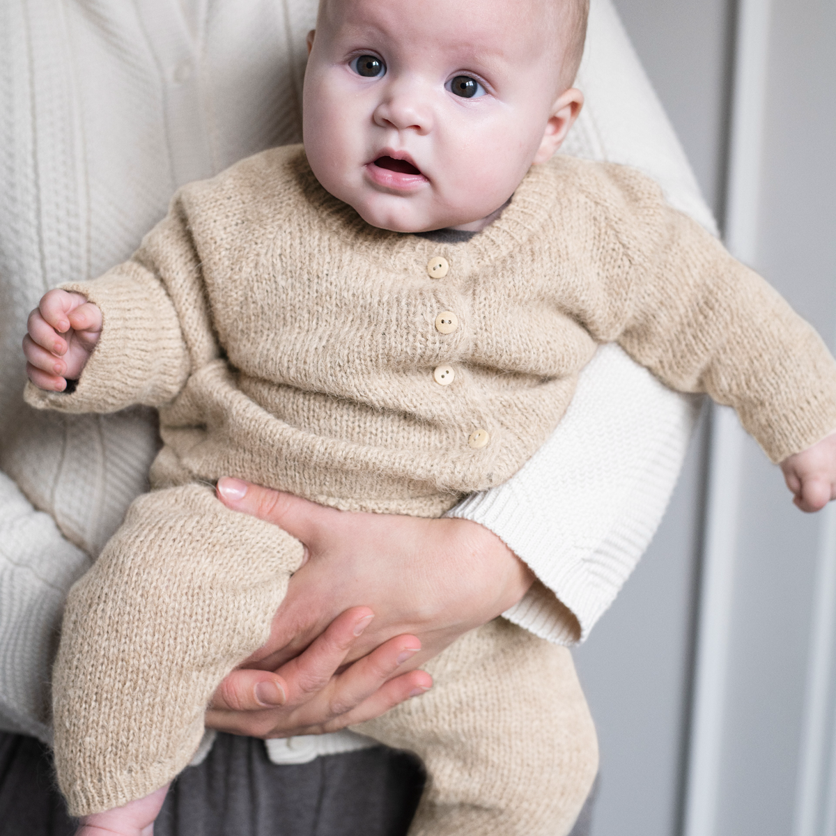 A baby wearing a beige knit cardigan and matching knit pants, held by an adult wearing a cream ribbed sweater. The baby is looking directly at the camera with a neutral gray background.