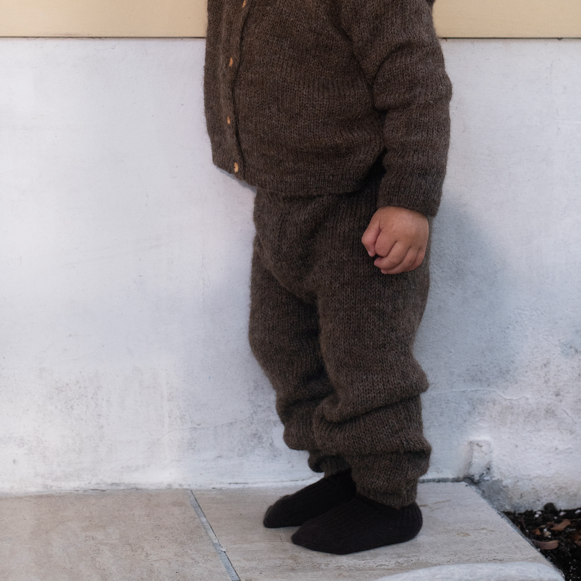 A toddler standing outdoors against a light wall, wearing a brown knit cardigan and matching pants with black socks.