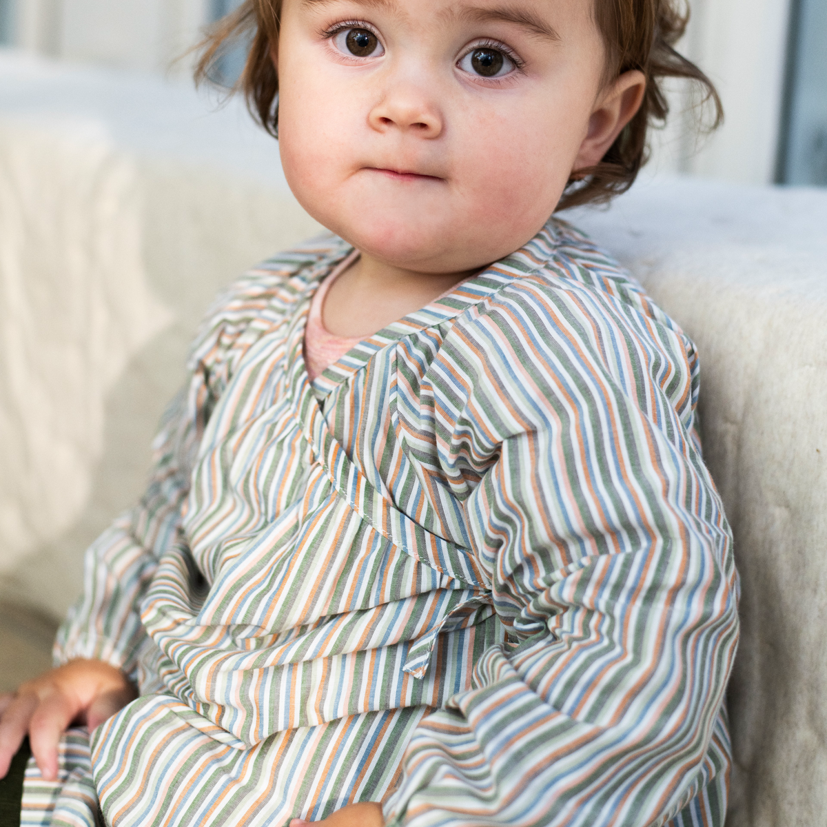 A close-up of a toddler wearing a long-sleeve wrap-style romper with vertical stripes in muted green, blue, orange, and beige, sitting on a light-colored fabric surface.