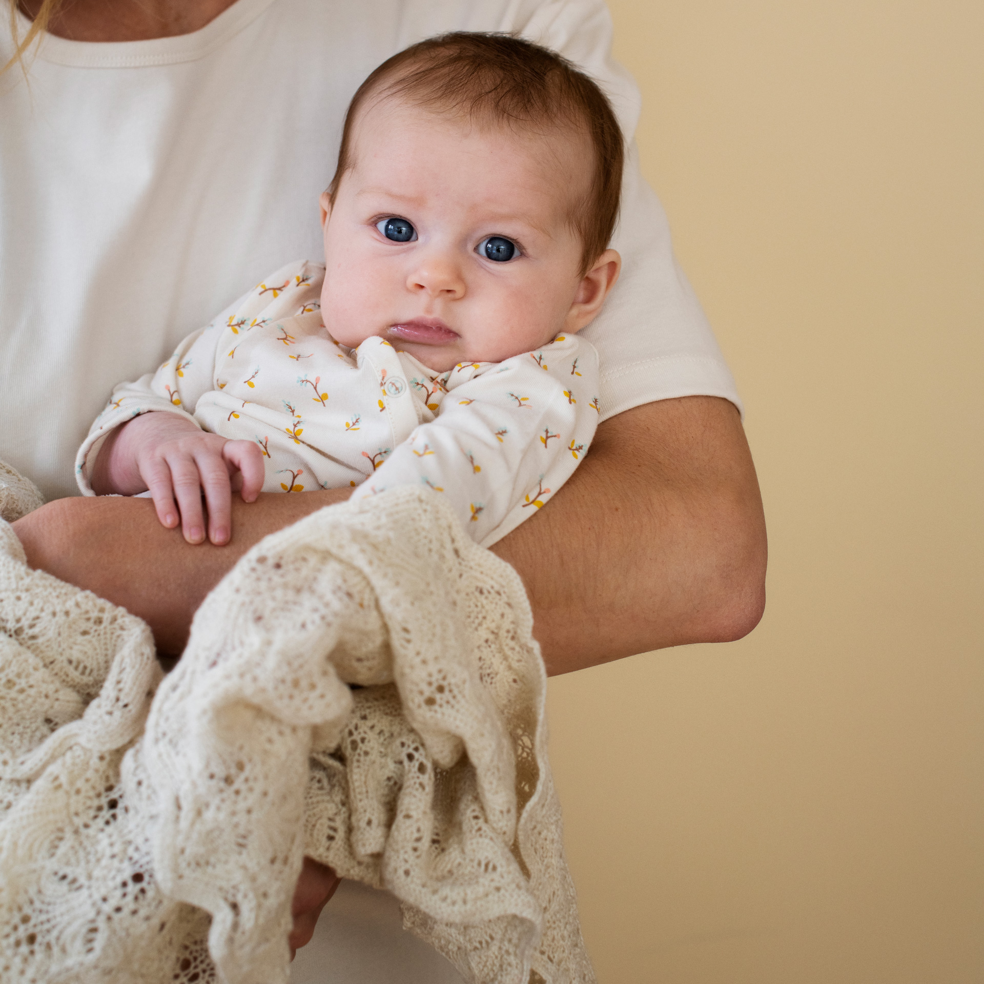 A baby wearing a white long-sleeve romper with tiny yellow motifs is held by an adult in a white top, wrapped in a cream-colored lace knit blanket against a solid beige background.