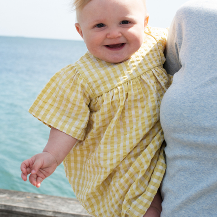 Baby in a yellow checkered dress standing by a body of water