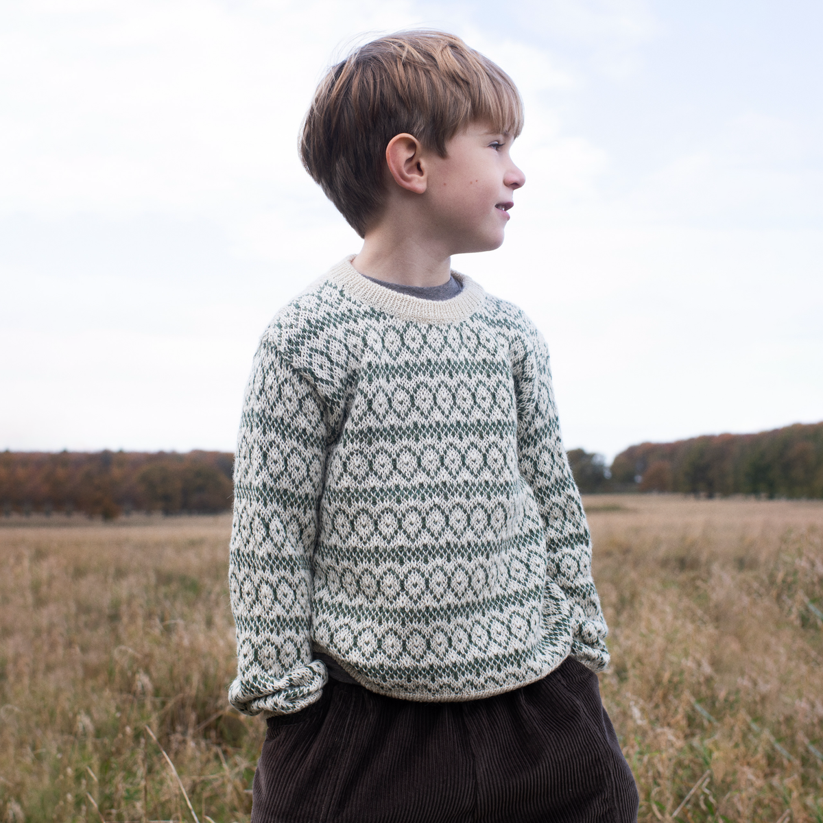 A young boy standing in a grassy field, wearing a cream and dark green patterned knit sweater and dark brown corduroy pants, looking off to the side.