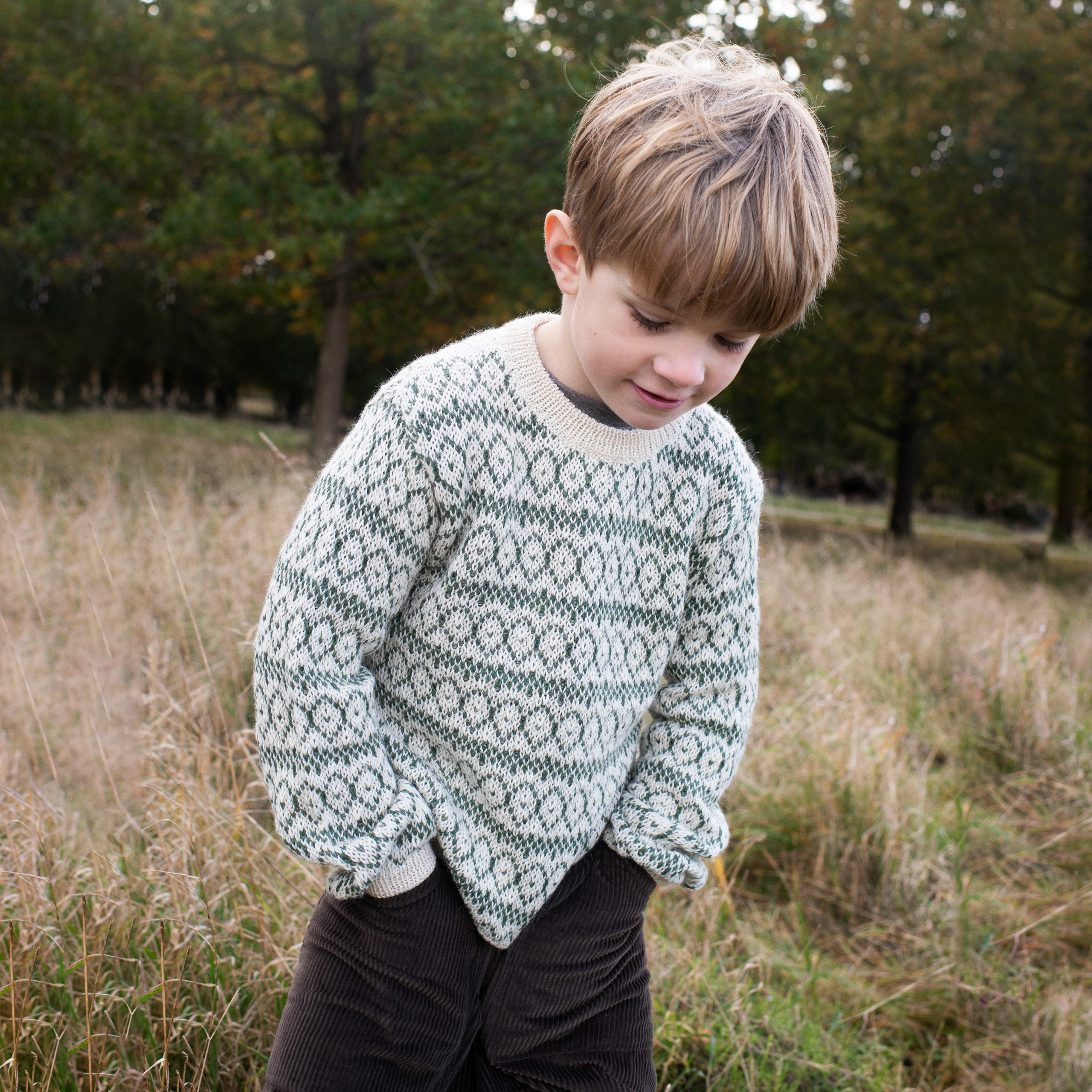 A young boy wearing a cream and dark green patterned knit sweater and dark corduroy pants, standing in a grassy field with trees in the background.
