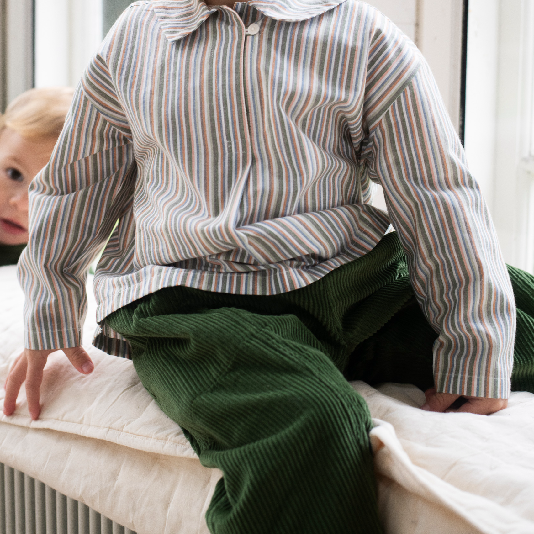 A child wearing a long-sleeve button-up shirt with thin vertical stripes in beige, brown, and muted green, paired with dark green corduroy pants, sitting on a white cushioned surface.