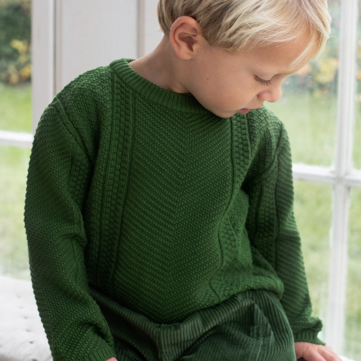 A young child with blonde hair wearing a dark green textured knit sweater and matching pants, sitting in front of a bright window.