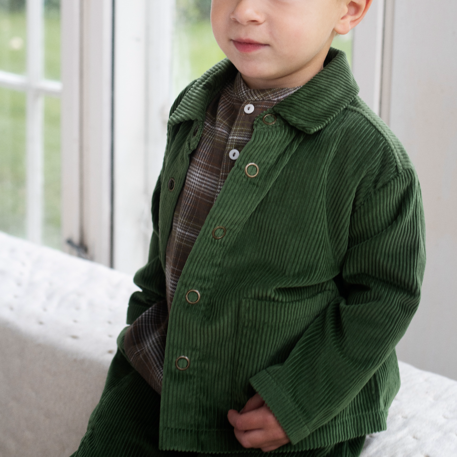 A toddler wearing a dark green corduroy jacket over a brown plaid shirt, sitting on a white tufted sofa in front of a window.