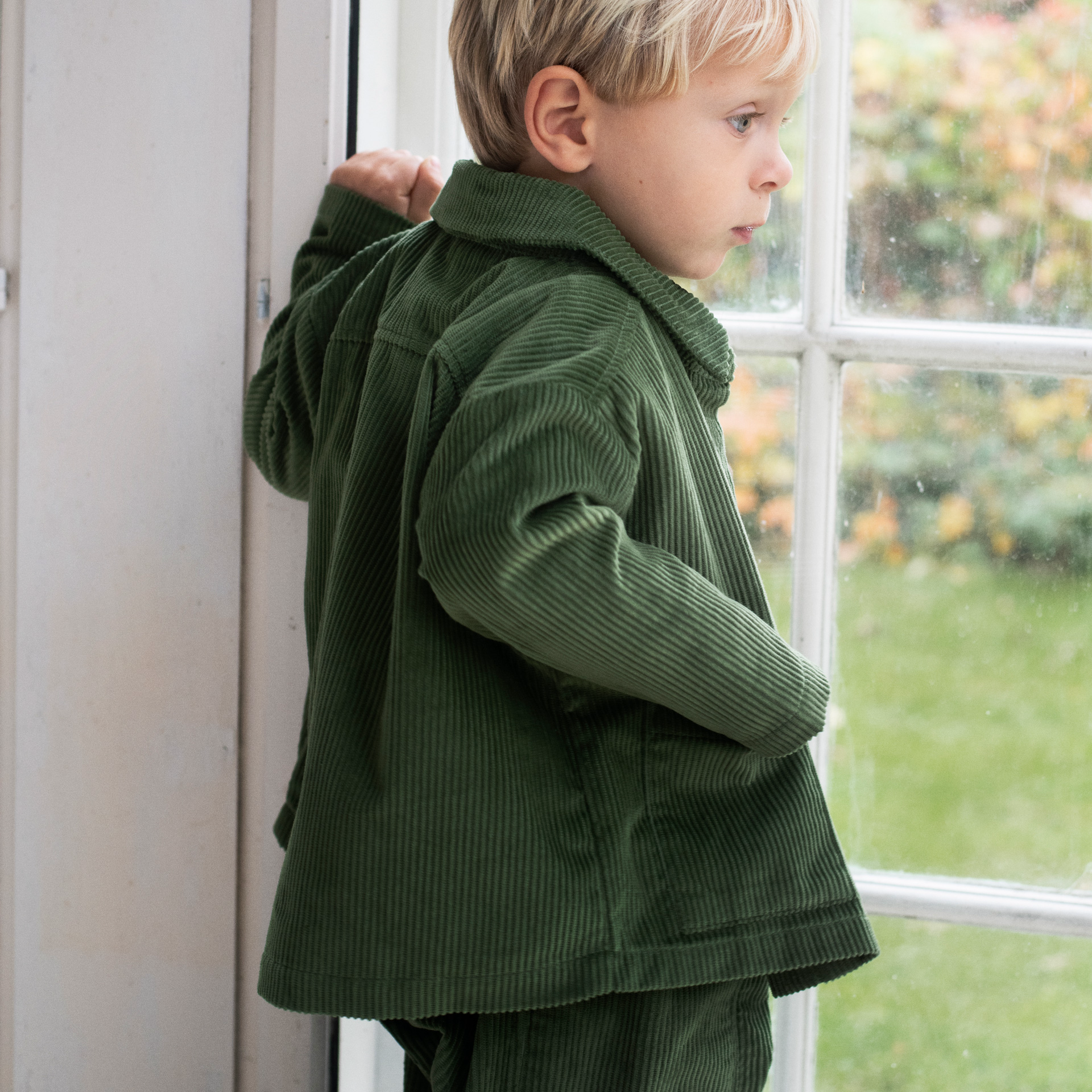 A young boy wearing a green corduroy jacket and matching trousers stands by a window, looking out onto a green garden.