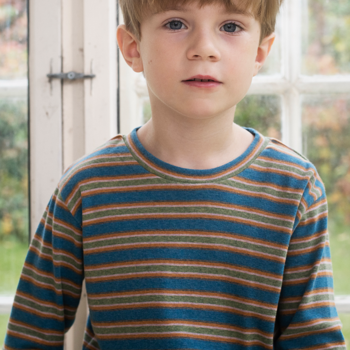 A young boy wearing a long-sleeve crewneck shirt with horizontal stripes in blue, green, and orange, standing in front of a white-framed window.