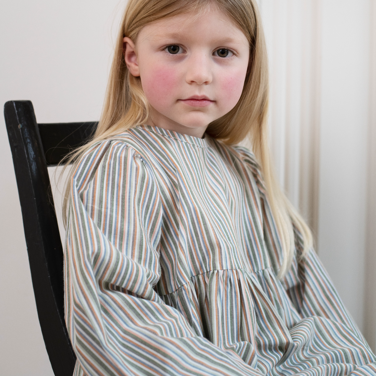 A young girl with long blonde hair sits on a black wooden chair, wearing a long-sleeved, high-neck dress with vertical stripes in muted blue, beige, and brown tones.