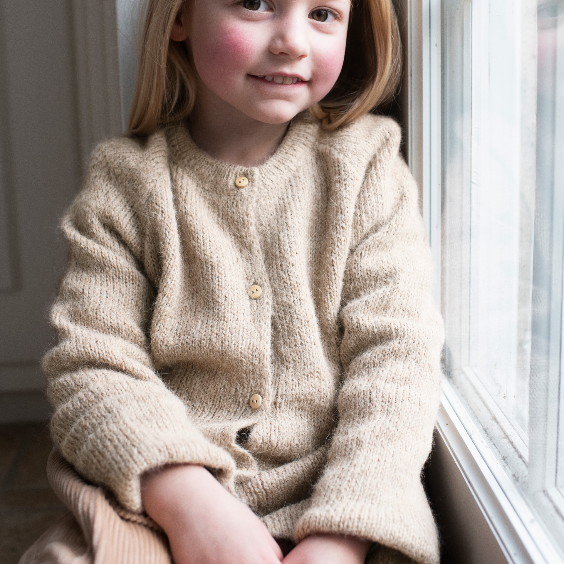 A young girl with blonde hair sits by a window wearing a cozy, knit beige cardigan with small wooden buttons.
