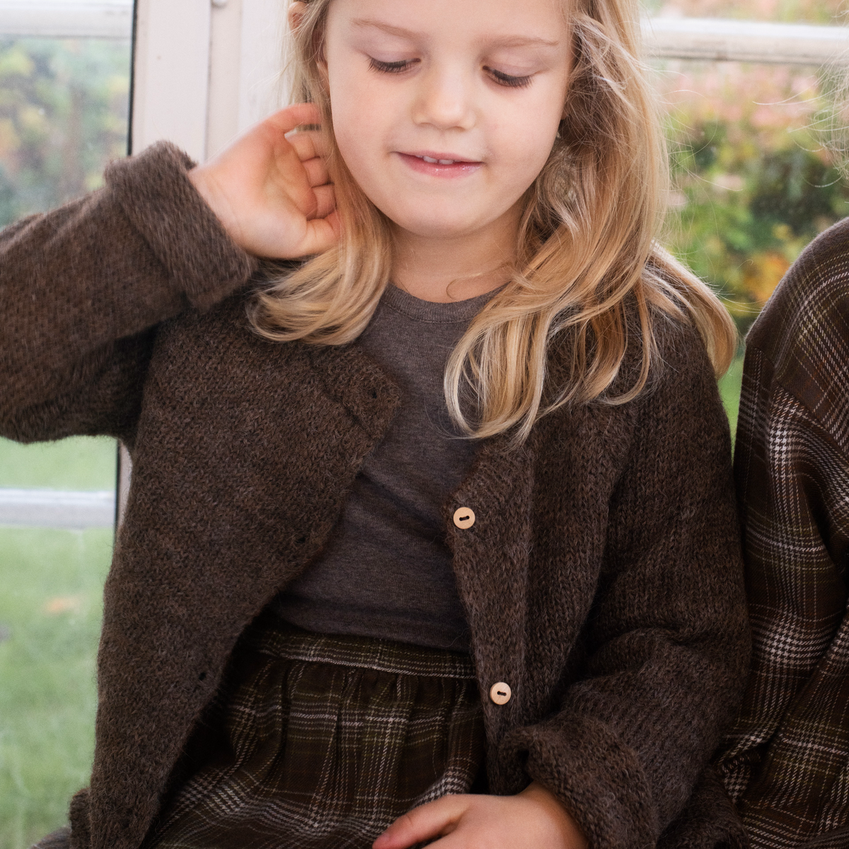 A young girl with blonde hair wearing a brown knit cardigan over a dark gray t-shirt and a brown plaid skirt, sitting indoors near a window.