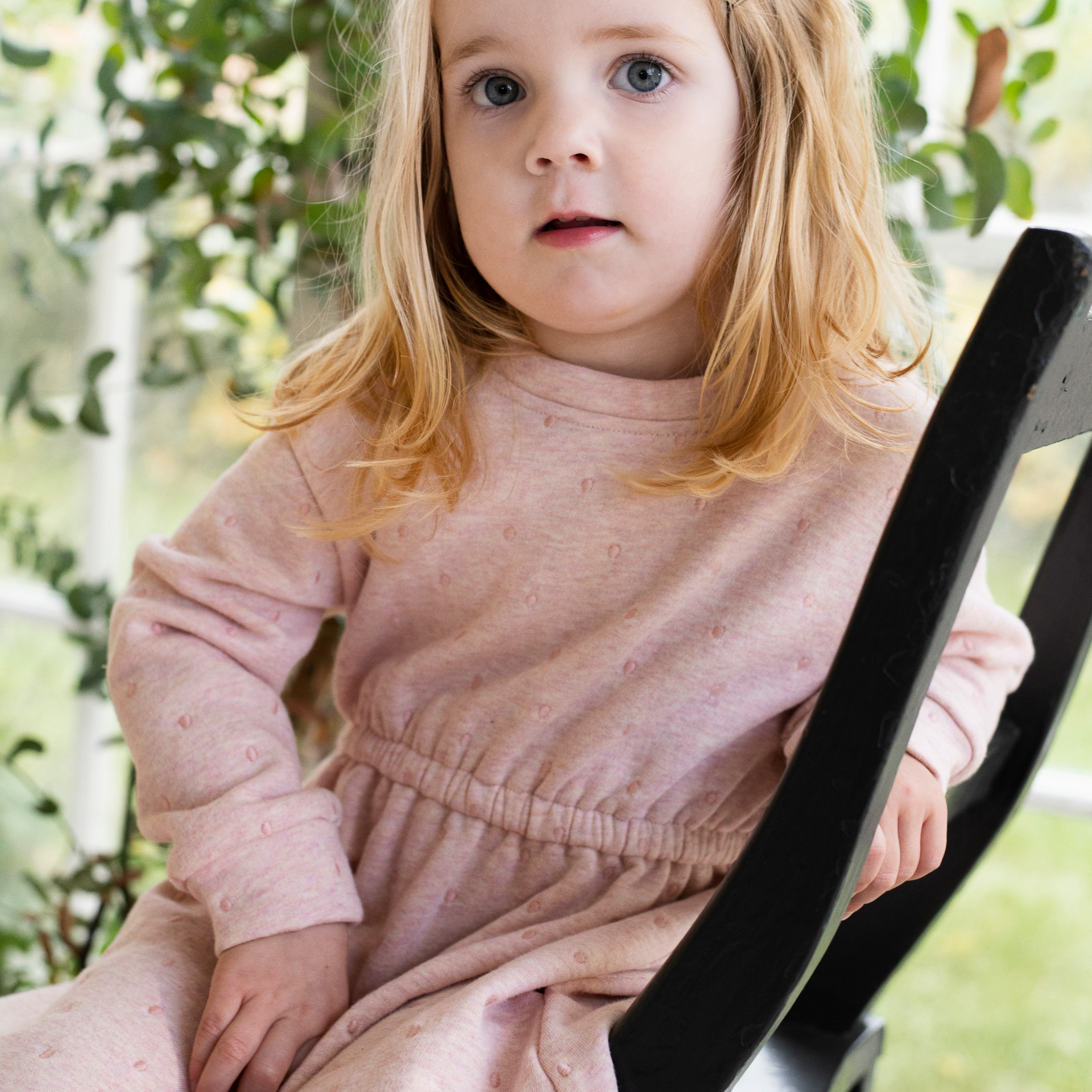 A young child with blonde hair wearing a dusty pink long-sleeve dress with tiny embroidered details, sitting on a black wooden chair in a garden setting.