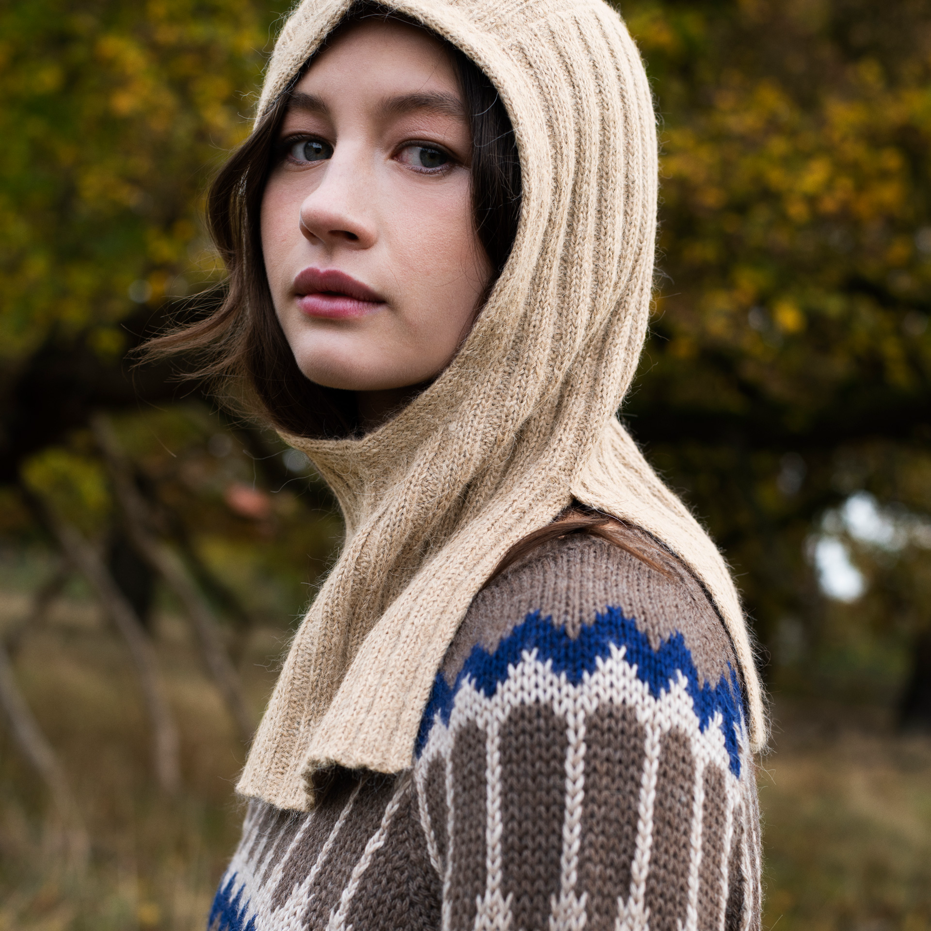 A woman wearing a beige ribbed knit hooded neck warmer over a brown, cream, and blue patterned knit sweater, photographed outdoors with a blurred autumn tree background.