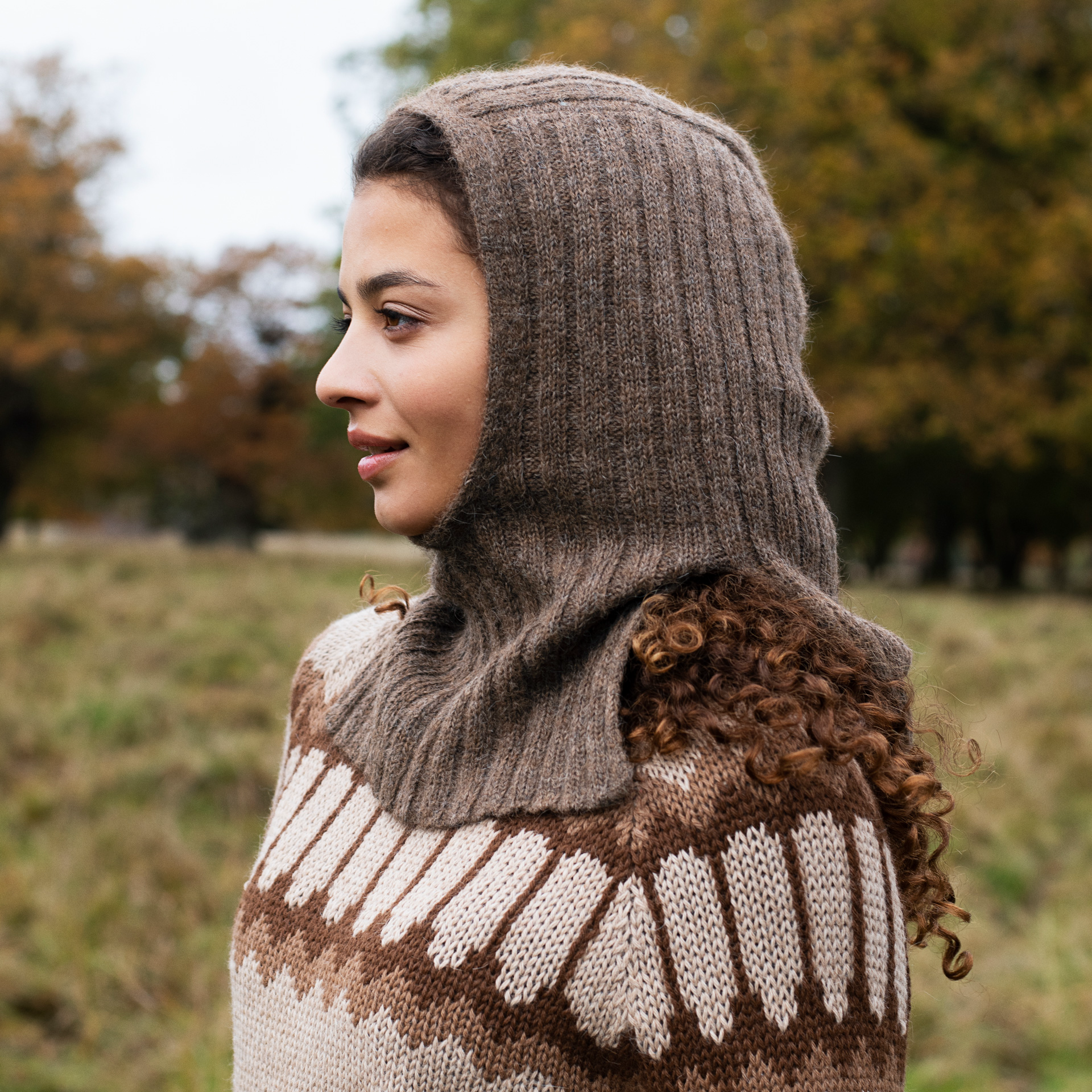 A woman wearing a brown ribbed knit hooded neck warmer over a brown and cream patterned knit sweater, photographed outdoors with a blurred autumn tree background.