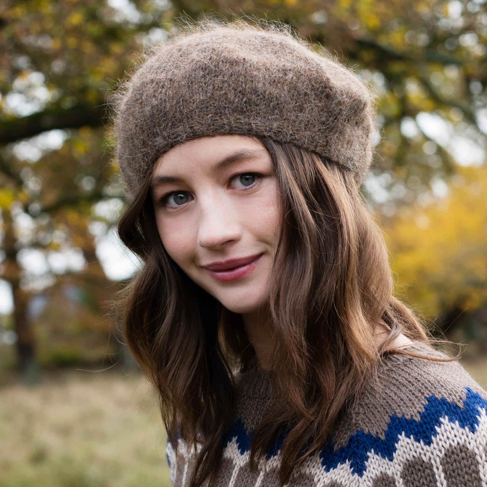 A woman wearing a brown knit beret and a brown, blue, and white patterned knit sweater, photographed outdoors with a blurred background of trees and grass.