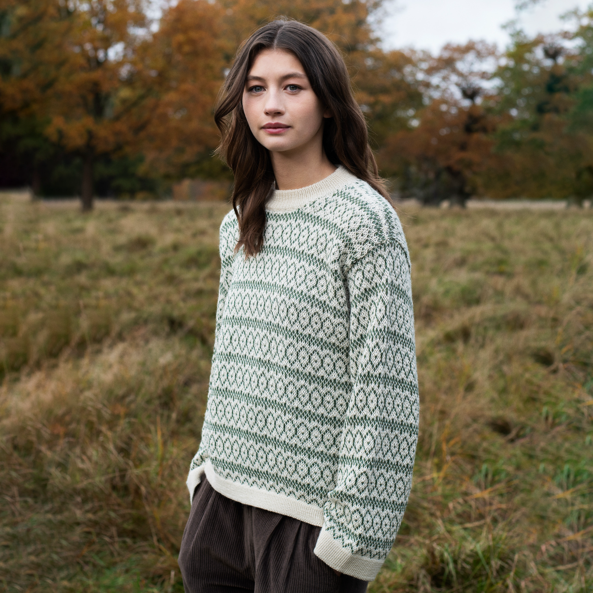 A woman wearing a cream and green patterned knit sweater stands in an outdoor grassy field with autumn trees in the background.
