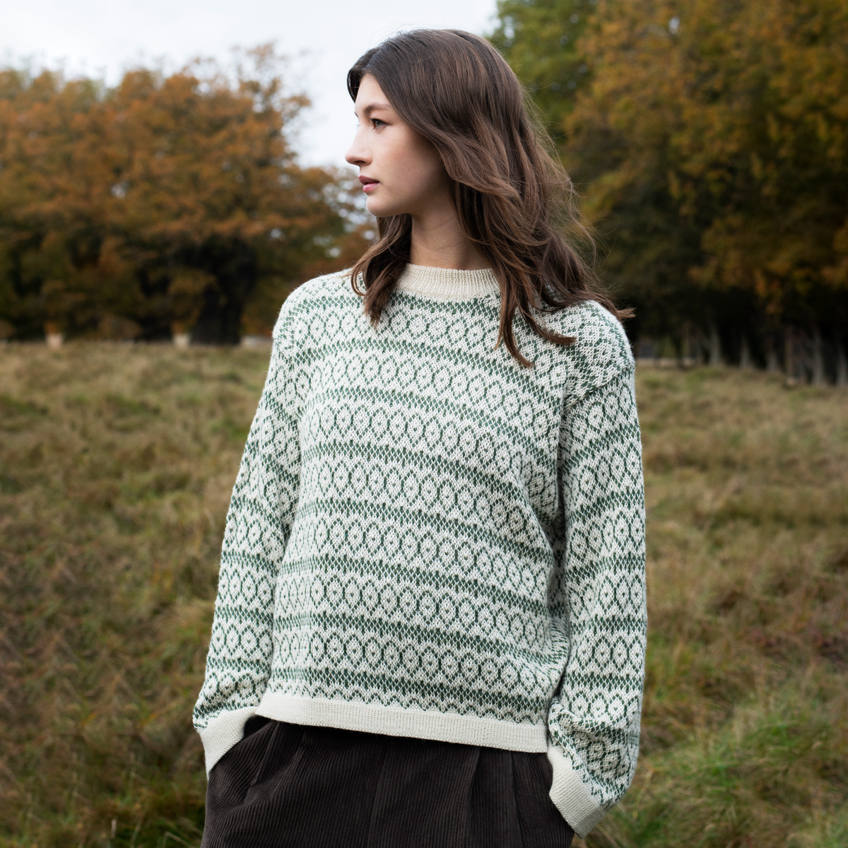 A woman wearing a cream and green patterned knit sweater stands outdoors in a grassy field with trees in the background.