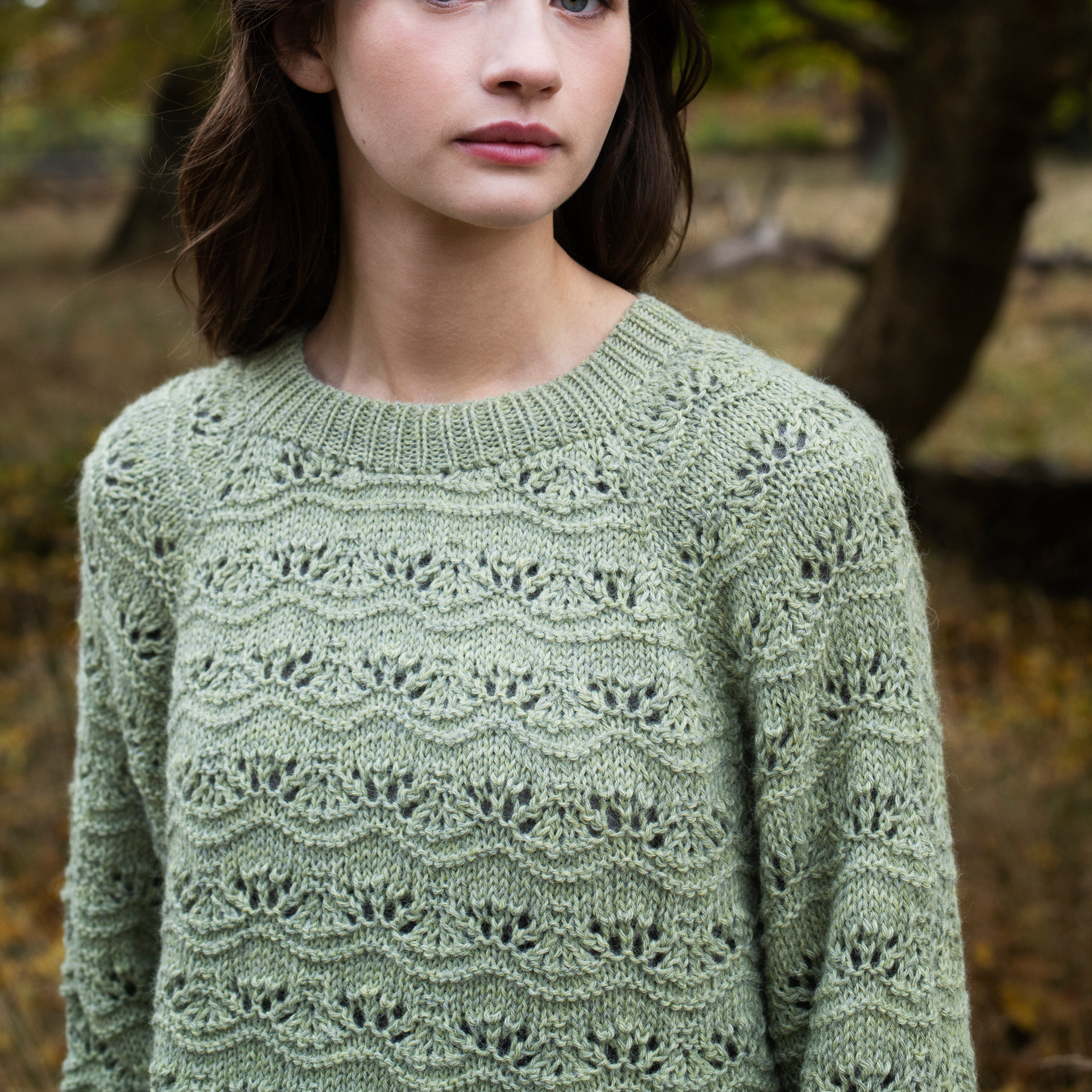 A woman wearing a light sage green knit sweater with an intricate leafy lace pattern, photographed outdoors in a softly blurred autumn park setting.