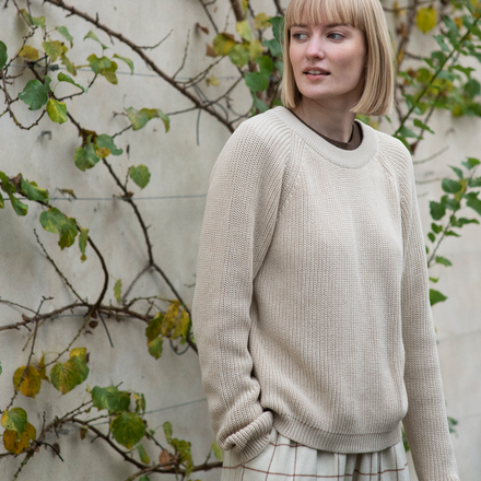 Woman wearing a beige sweater standing in front of a wall with greenery