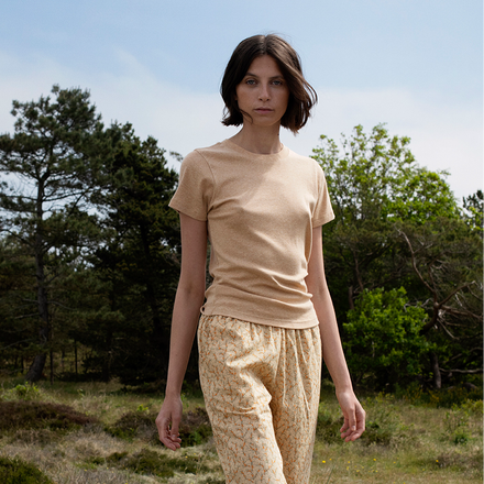 Woman in orange top and pants standing in a natural setting with trees and grass.