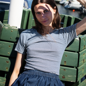 Woman in a striped shirt and blue skirt sitting on green wooden crates by a waterfront.