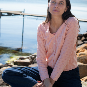 Woman wearing a pink sweater sitting on rocks by a body of water.