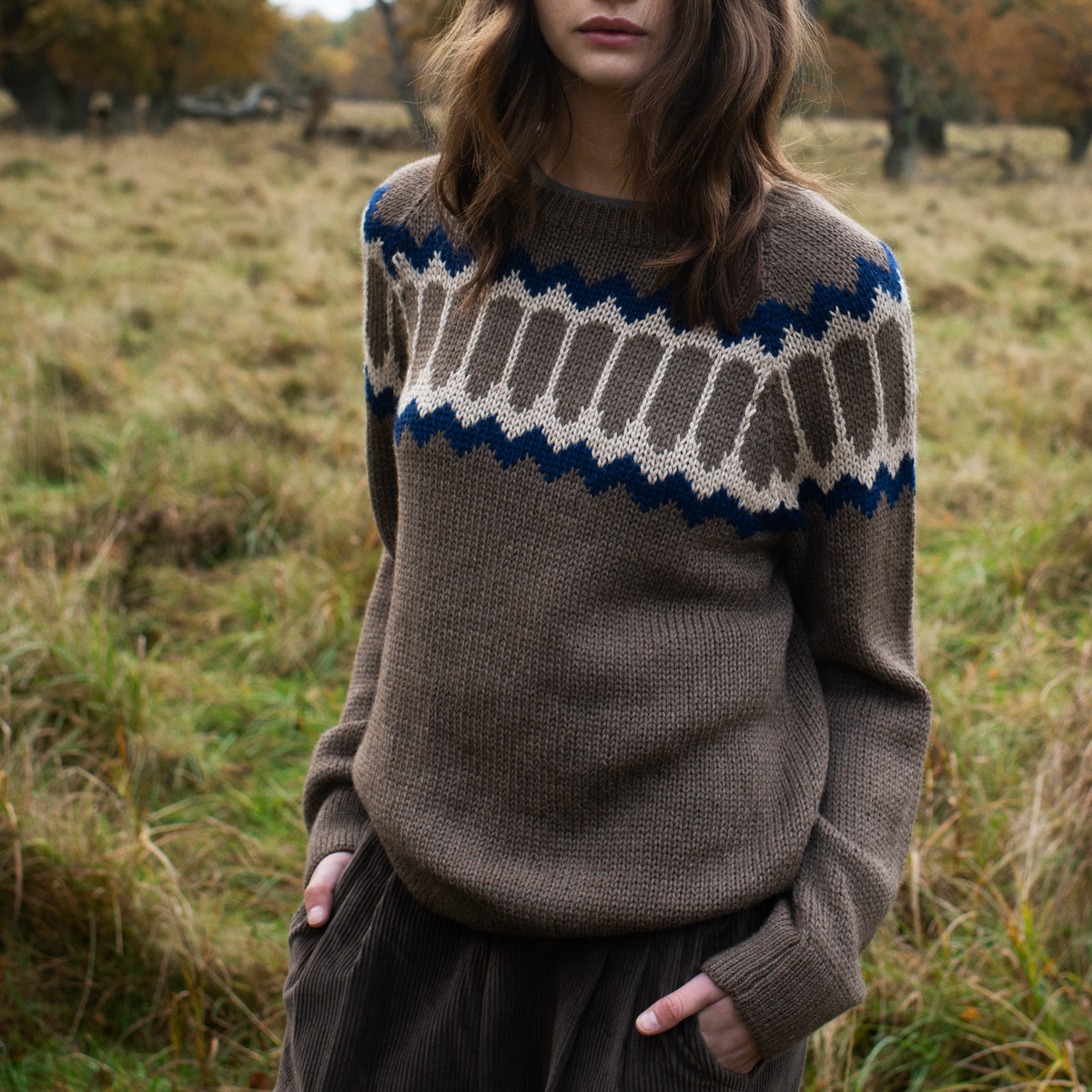 A woman wearing a brown knit sweater with a Fair Isle pattern across the chest in navy blue and beige, shown against a blurred autumn background.