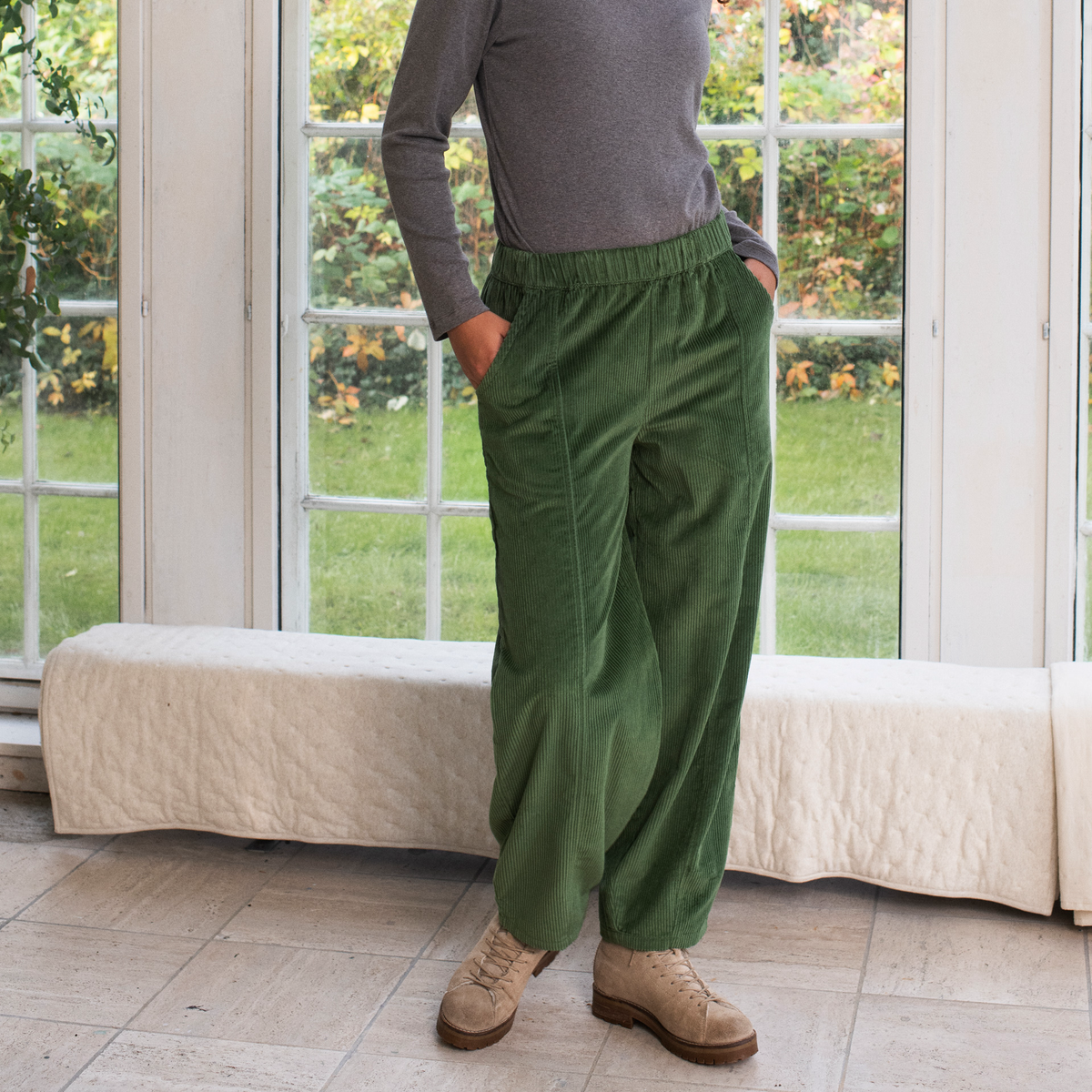 A person wearing loose, wide-leg olive green corduroy pants with an elastic waistband, paired with a gray long-sleeve top and beige lace-up shoes, standing in a bright room with large windows and a bench.