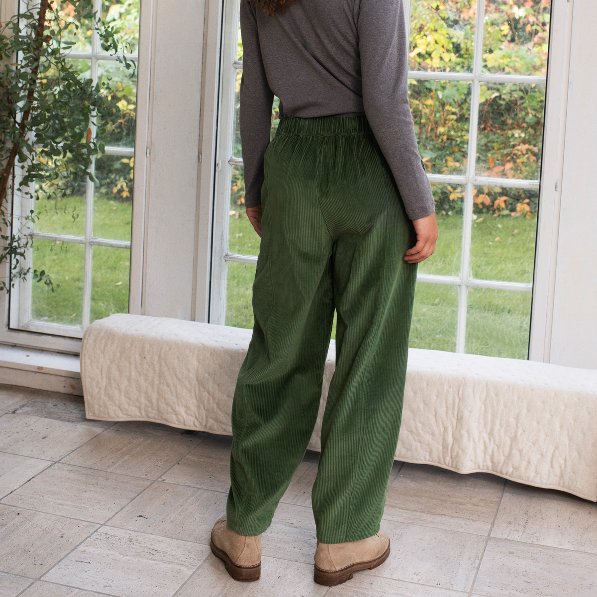 A person shown from the back wearing loose, wide-leg olive green corduroy trousers, a gray long-sleeve top, and tan boots, standing in a bright room with large windows and a white bench.