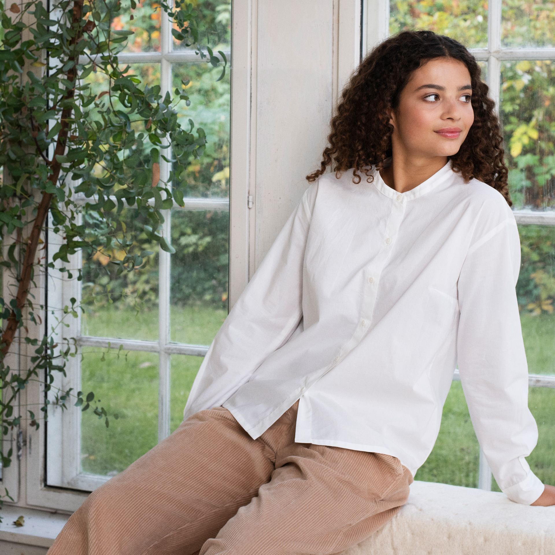 A woman with curly dark hair sits by a large white-framed window, wearing a white long-sleeve button-front shirt and beige trousers. A green plant is visible on the left, and the outdoor yard appears through the window.