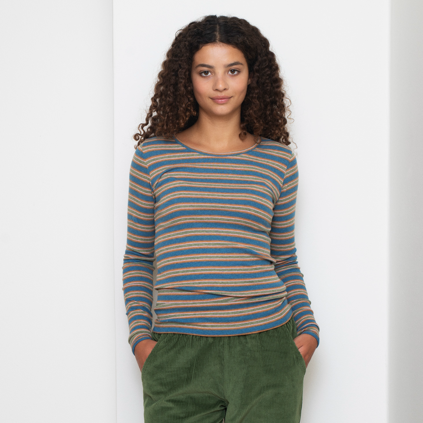 A woman with curly brown hair wearing a blue, tan, and brown striped long-sleeve knit top and olive green corduroy pants, standing against a white wall.