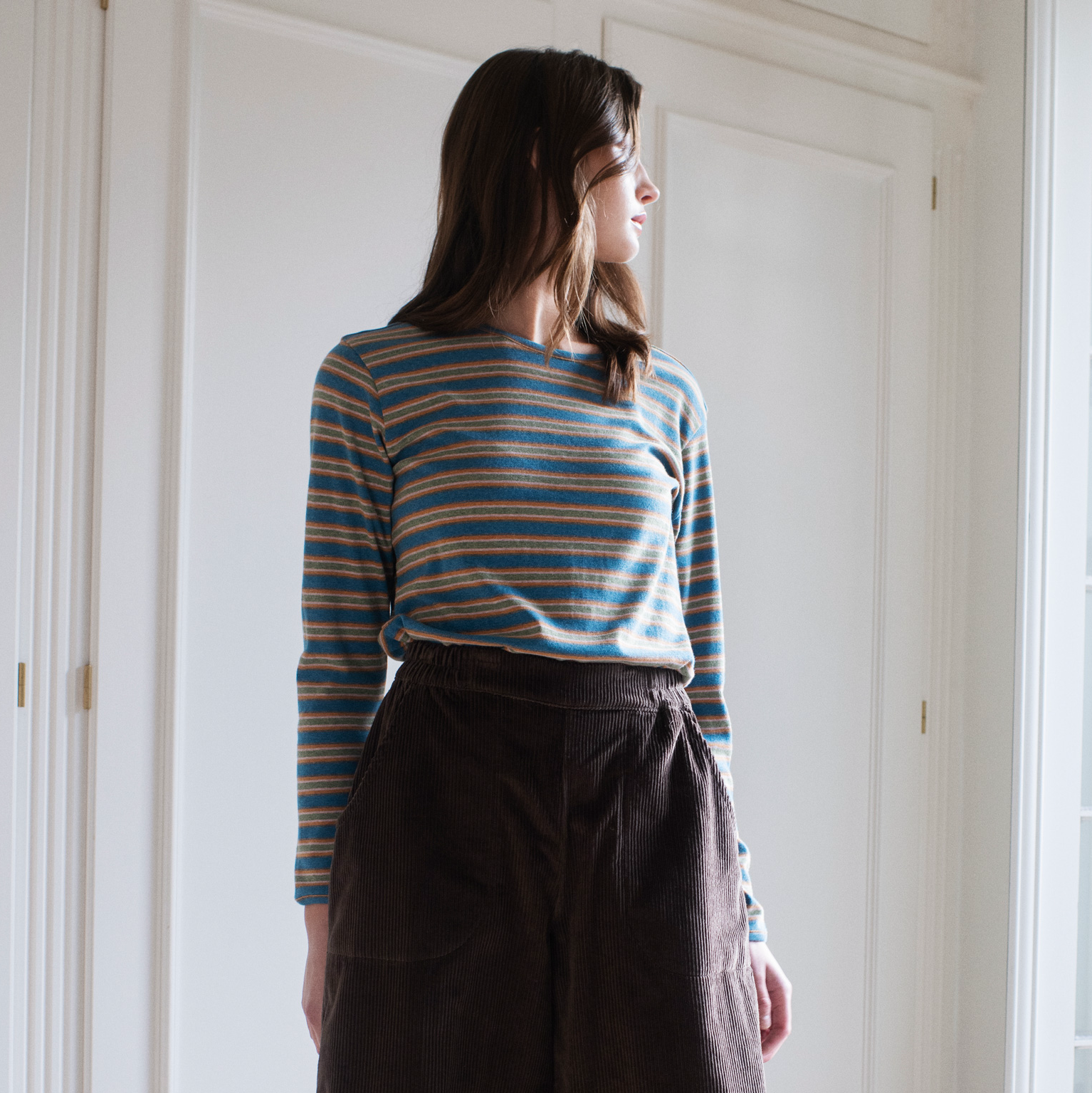 A woman wearing a blue, orange, and yellow striped long-sleeve knit top tucked into dark brown corduroy trousers, standing in a brightly lit room with white paneled doors.
