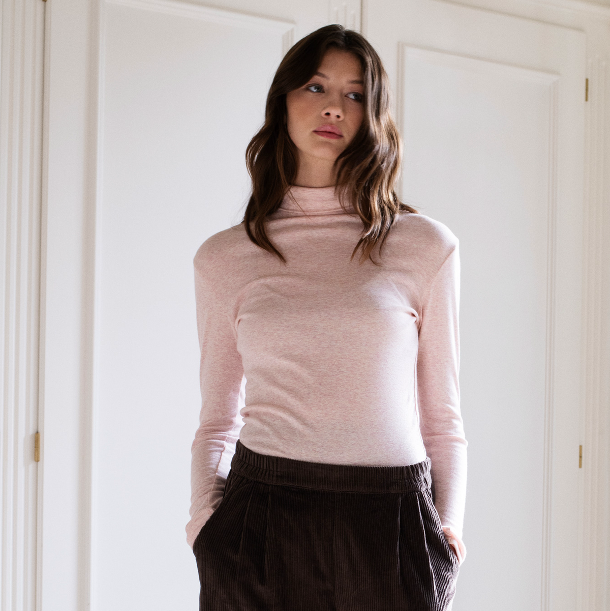 A woman wearing a light pink long-sleeve turtleneck top and dark brown corduroy trousers, standing in front of white paneled doors.