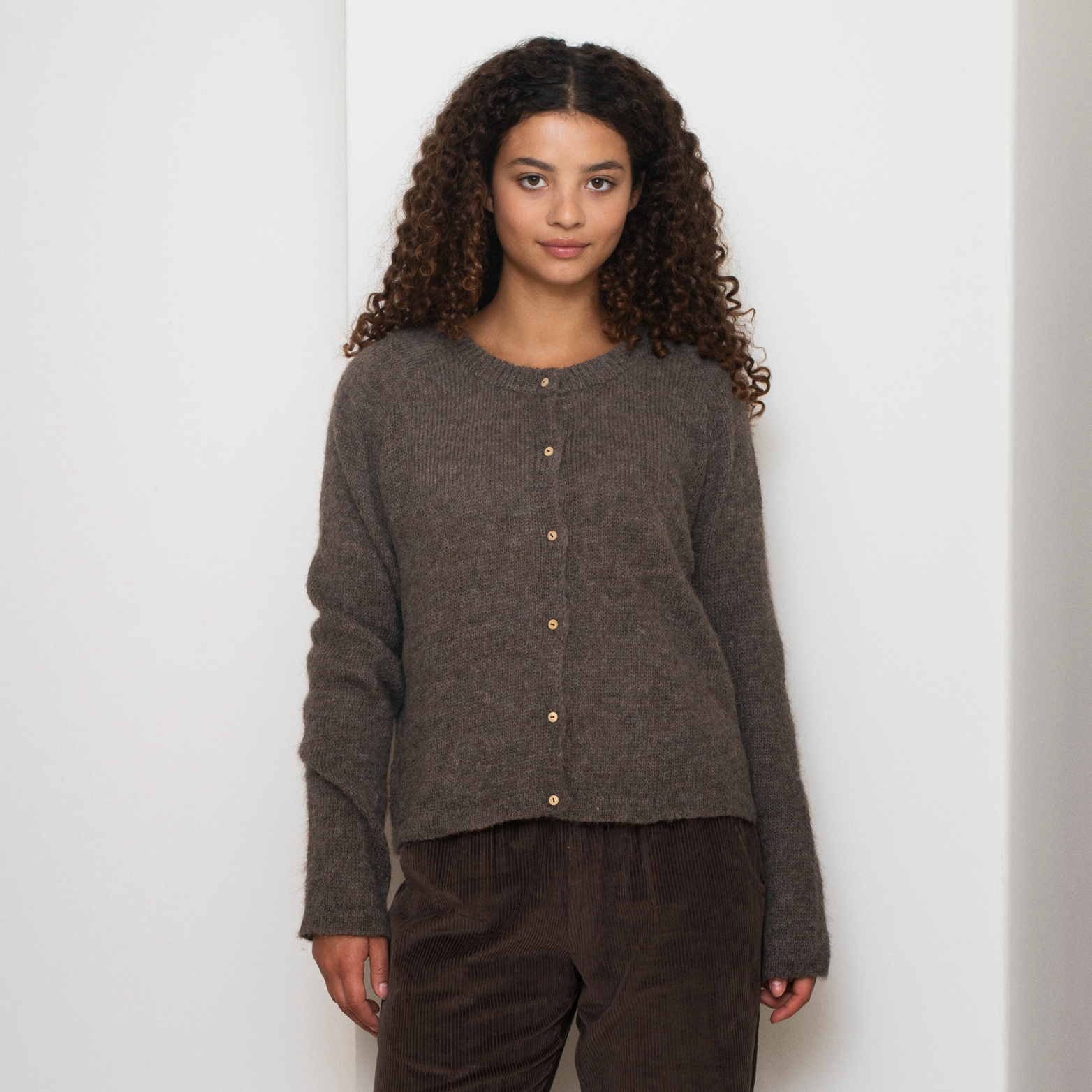 A woman with curly brown hair wearing a dark brown button-up knit sweater and dark brown corduroy pants, standing against a plain white wall.