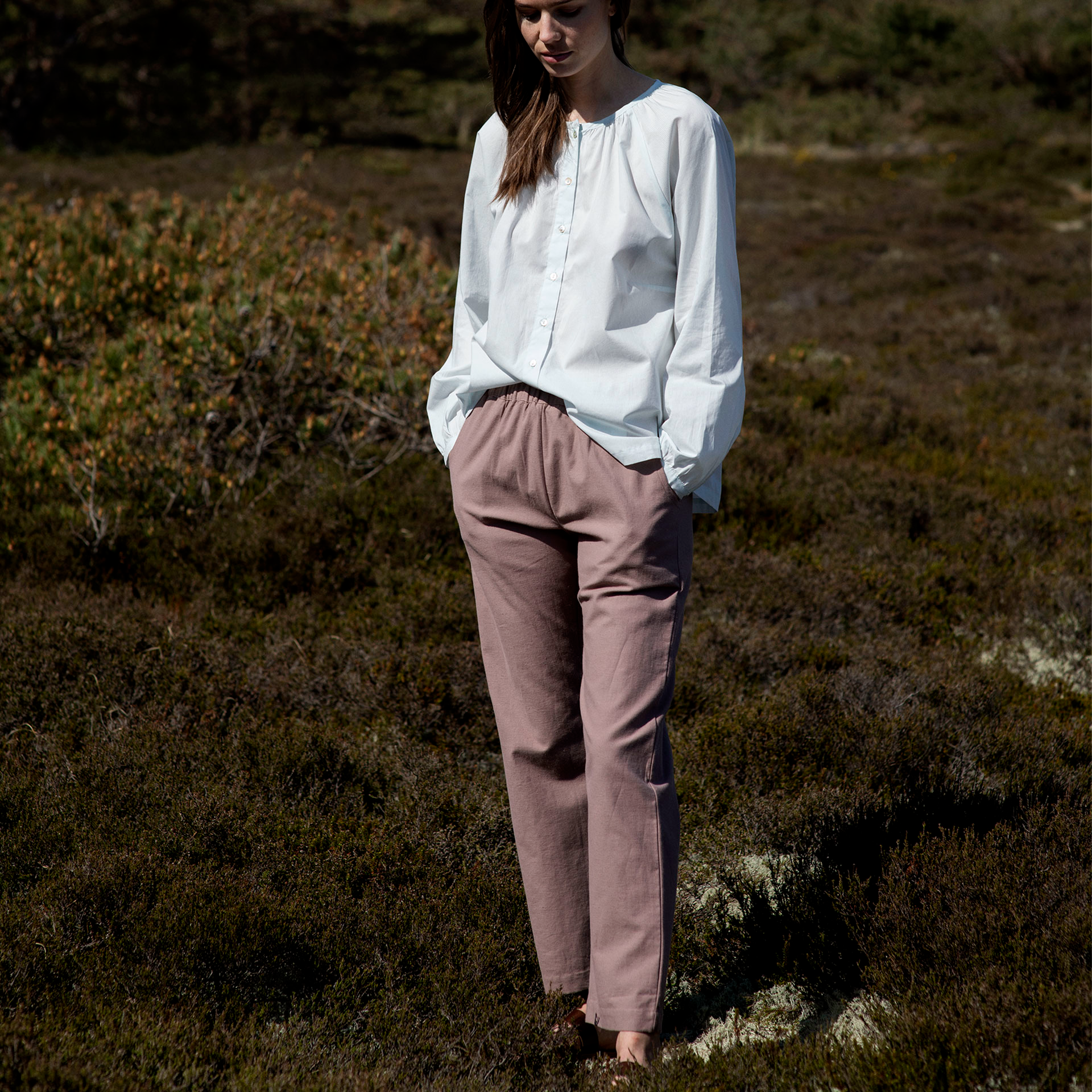 Woman in a white blouse and brown pants standing in a natural landscape with trees and grass.