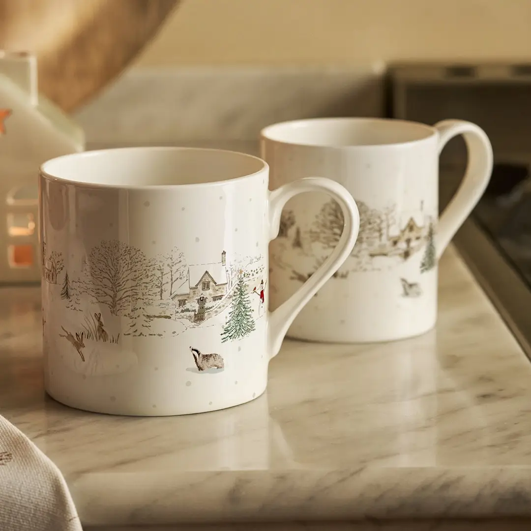 Two white china mugs with winter village illustrations, displayed on a marble kitchen countertop.