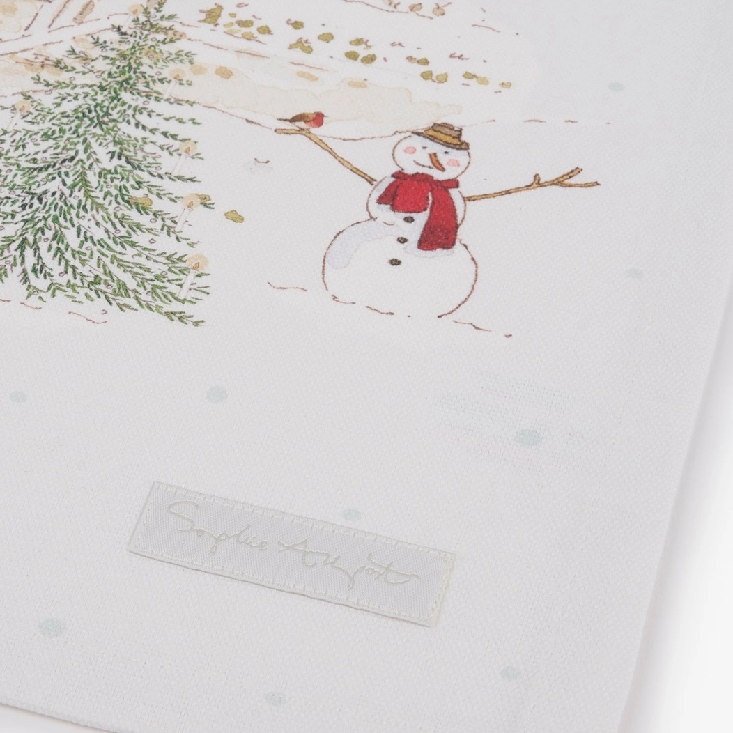 A close-up of a white tea towel, featuring a printed winter scene with a snowman, evergreen tree, and small bird. A small fabric label with the brand name Sophie Allport is visible near the bottom.