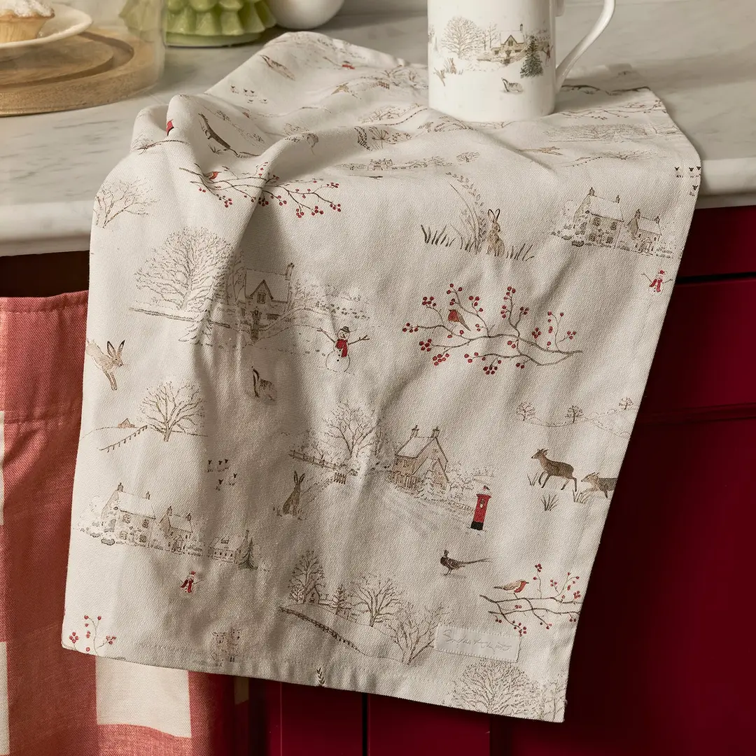 An off-white kitchen towel with a winter village print, draped over a dark red kitchen cabinet with a white mug resting on top.