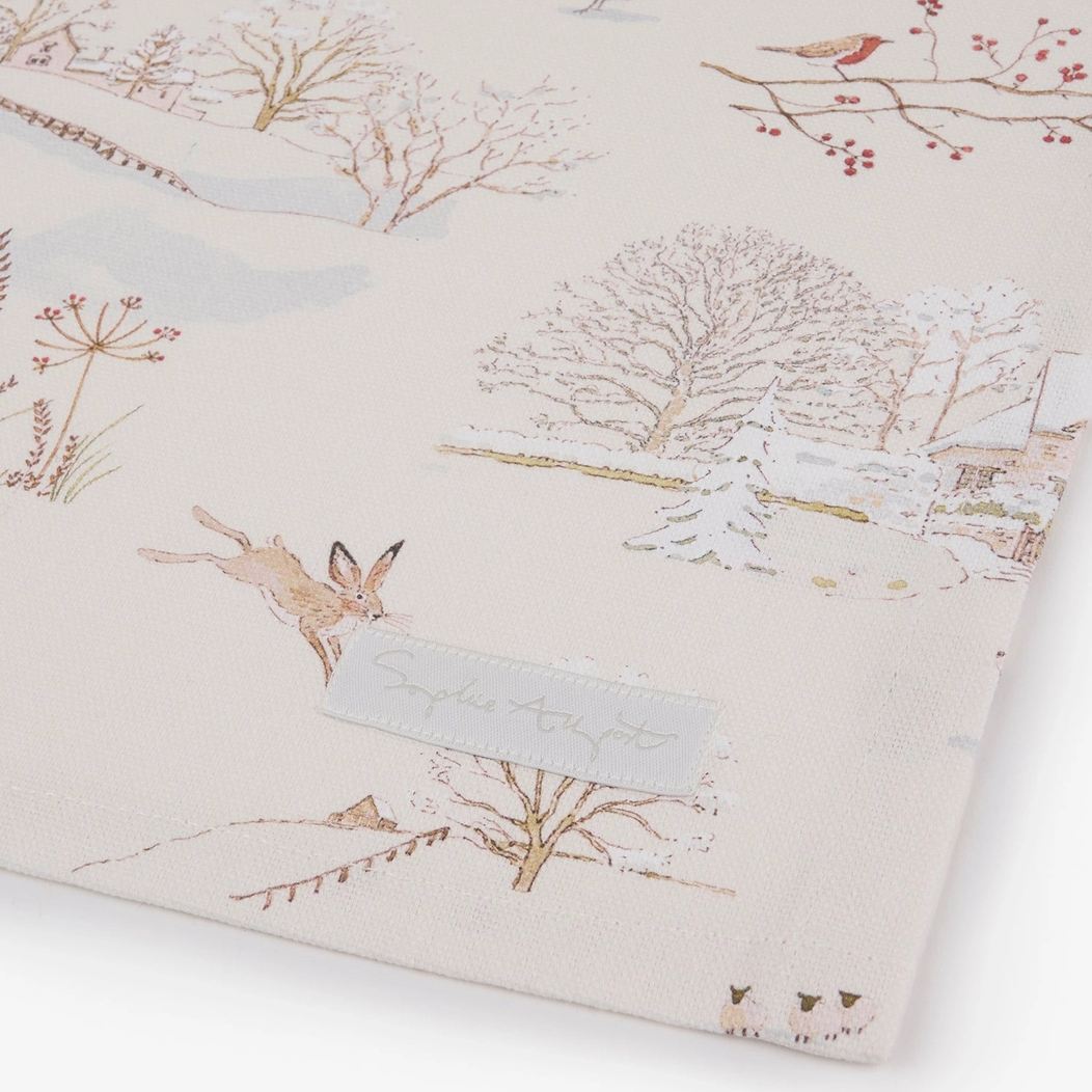 A close-up of a tea towel featuring a winter landscape print with trees, birds, and a hare on a cream background.
