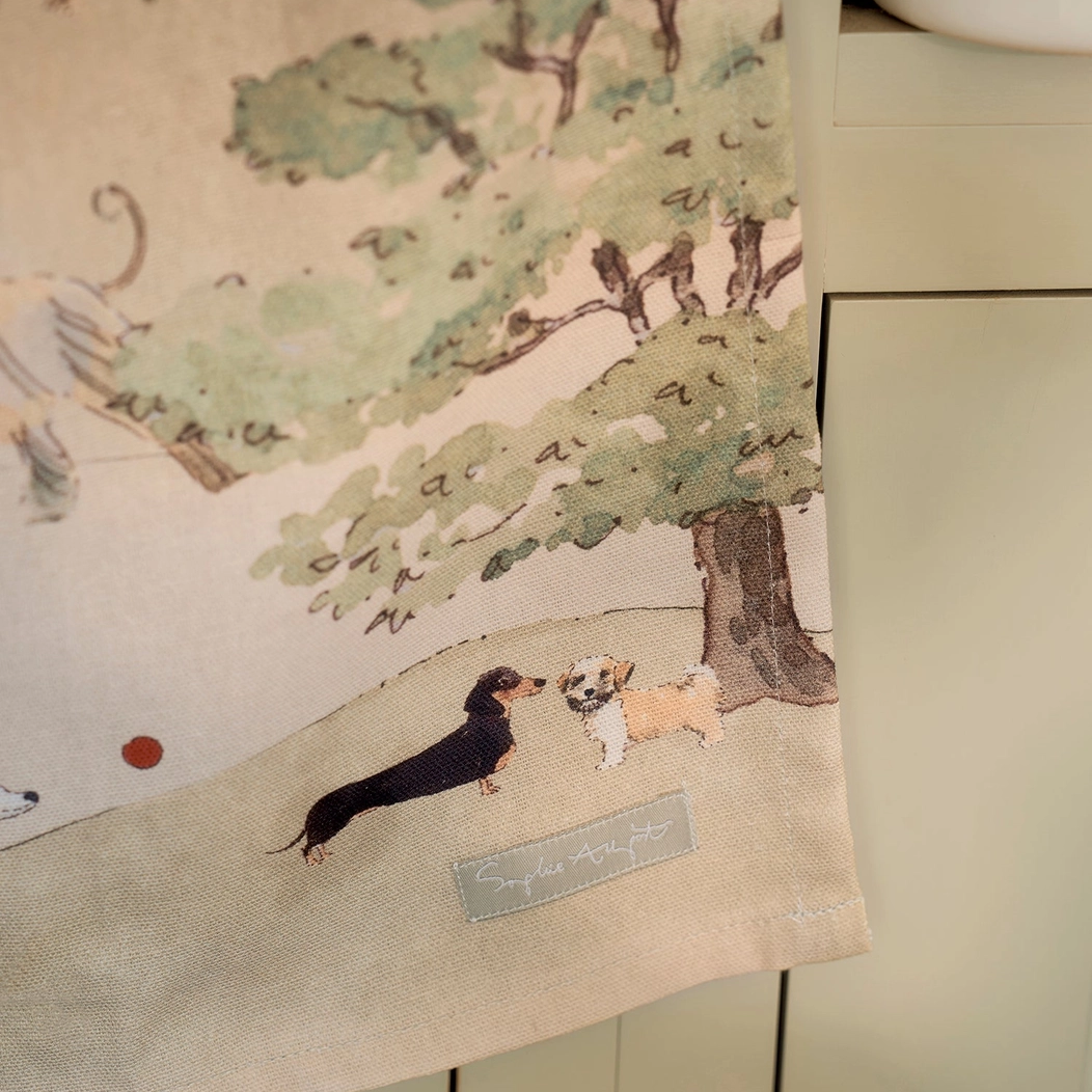 A close-up of a tea towel featuring a watercolor-style illustration of two dogs under a tree, hanging against a light-colored cabinet background.