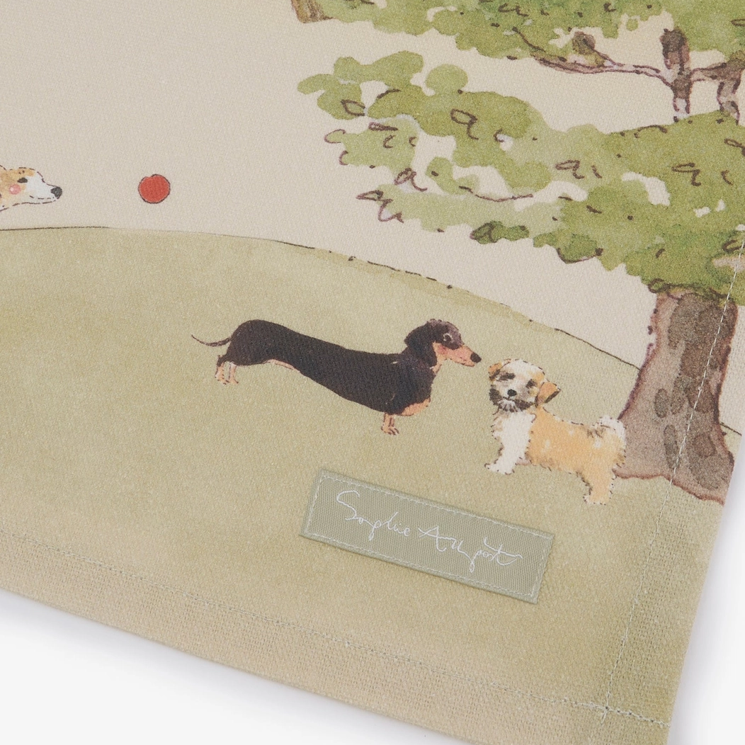 A close-up of a beige tea towel featuring a watercolor-style illustration of dachshunds in a park with a tree and a grey brand label.