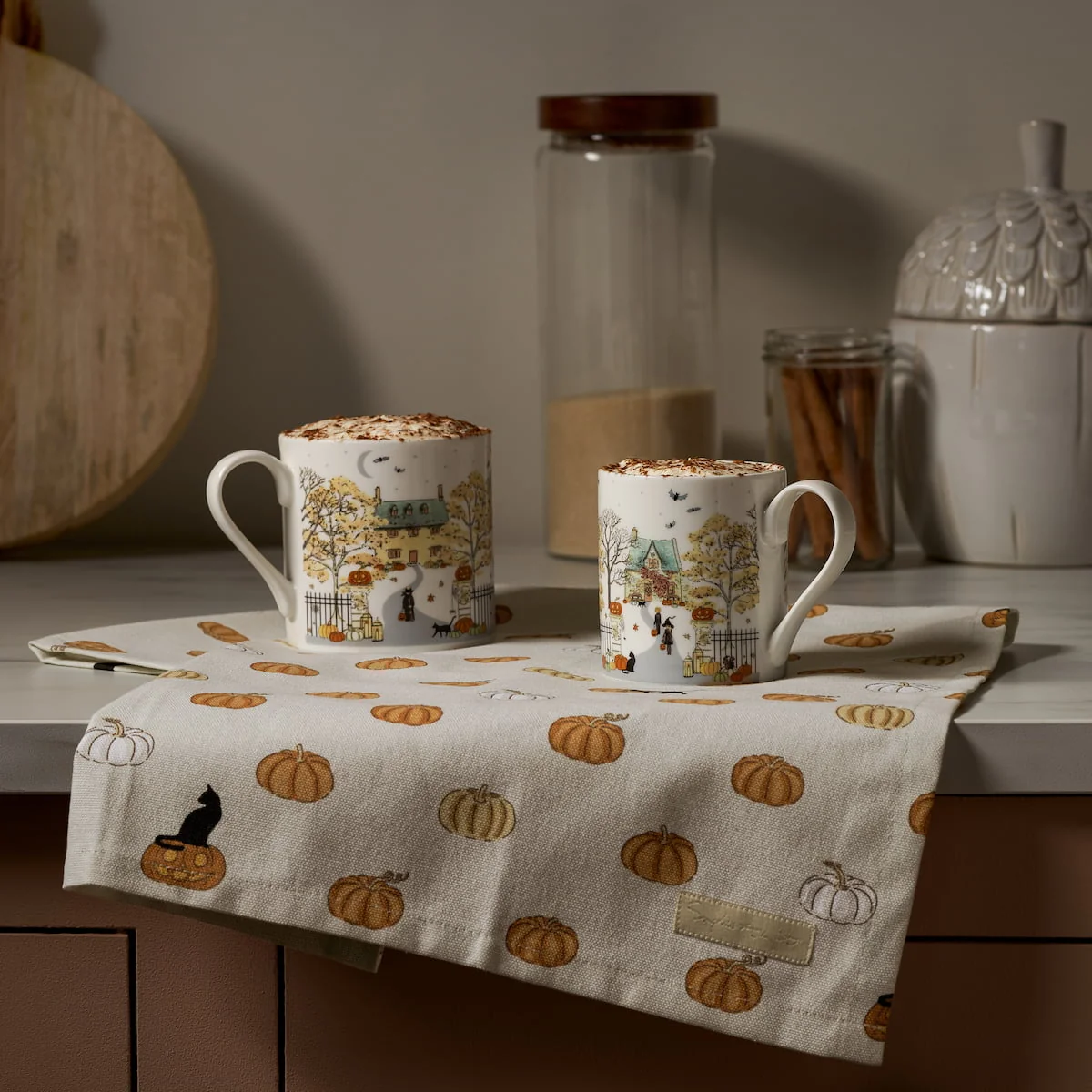 Two china mugs with Halloween scenes sitting atop a seasonal tea towel.